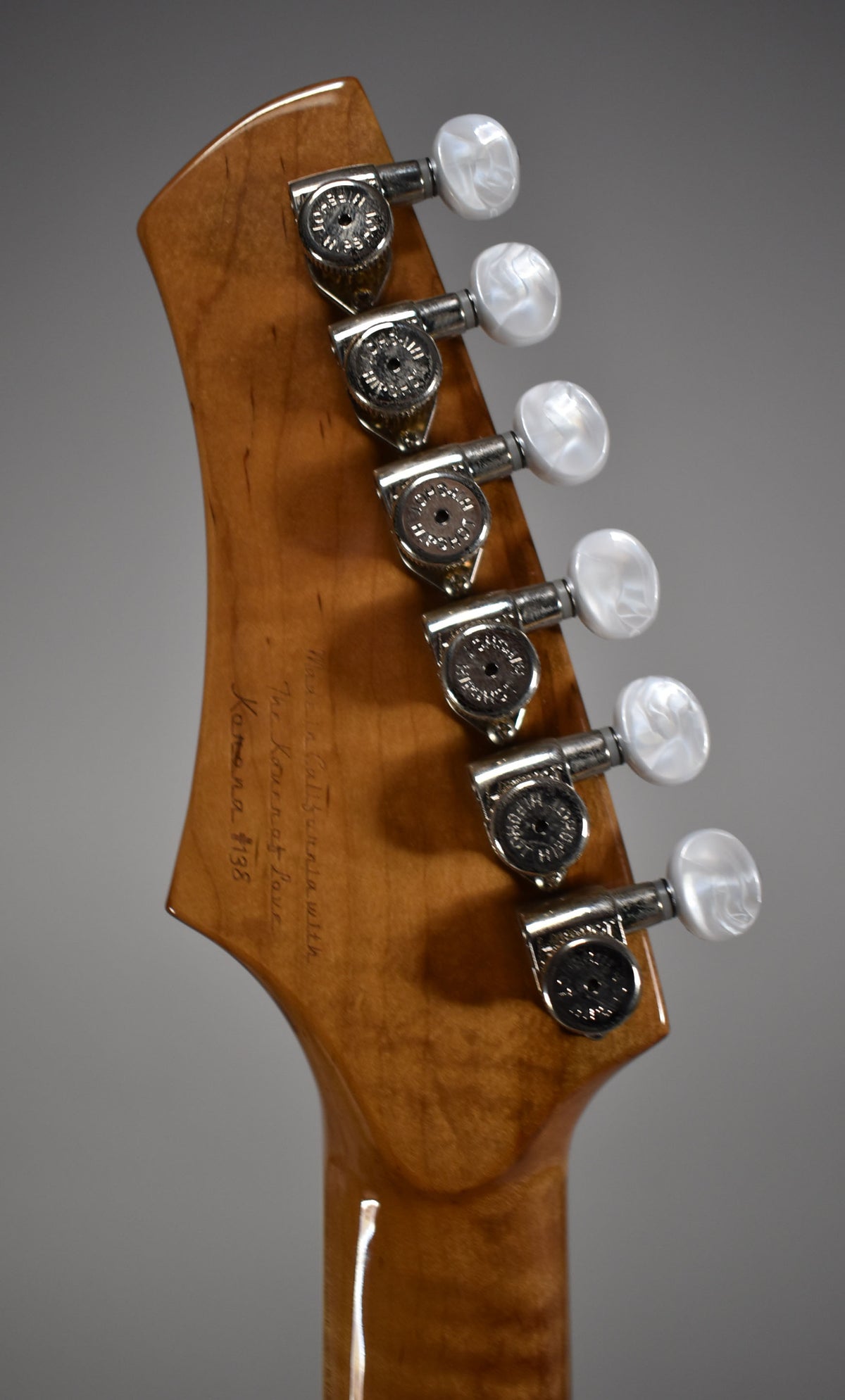 The back of a guitar headstock and neck in a glossy light brown finish, with six chrome tuners and pearloid buttons.