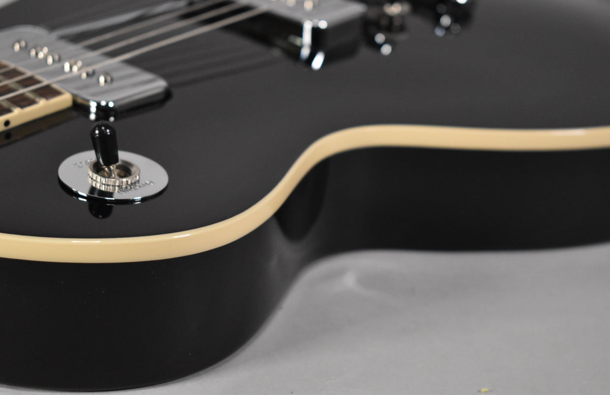 Close-up of the upper bout of a glossy black Les Paul, showing the cream binding and chrome pickup selector switch.