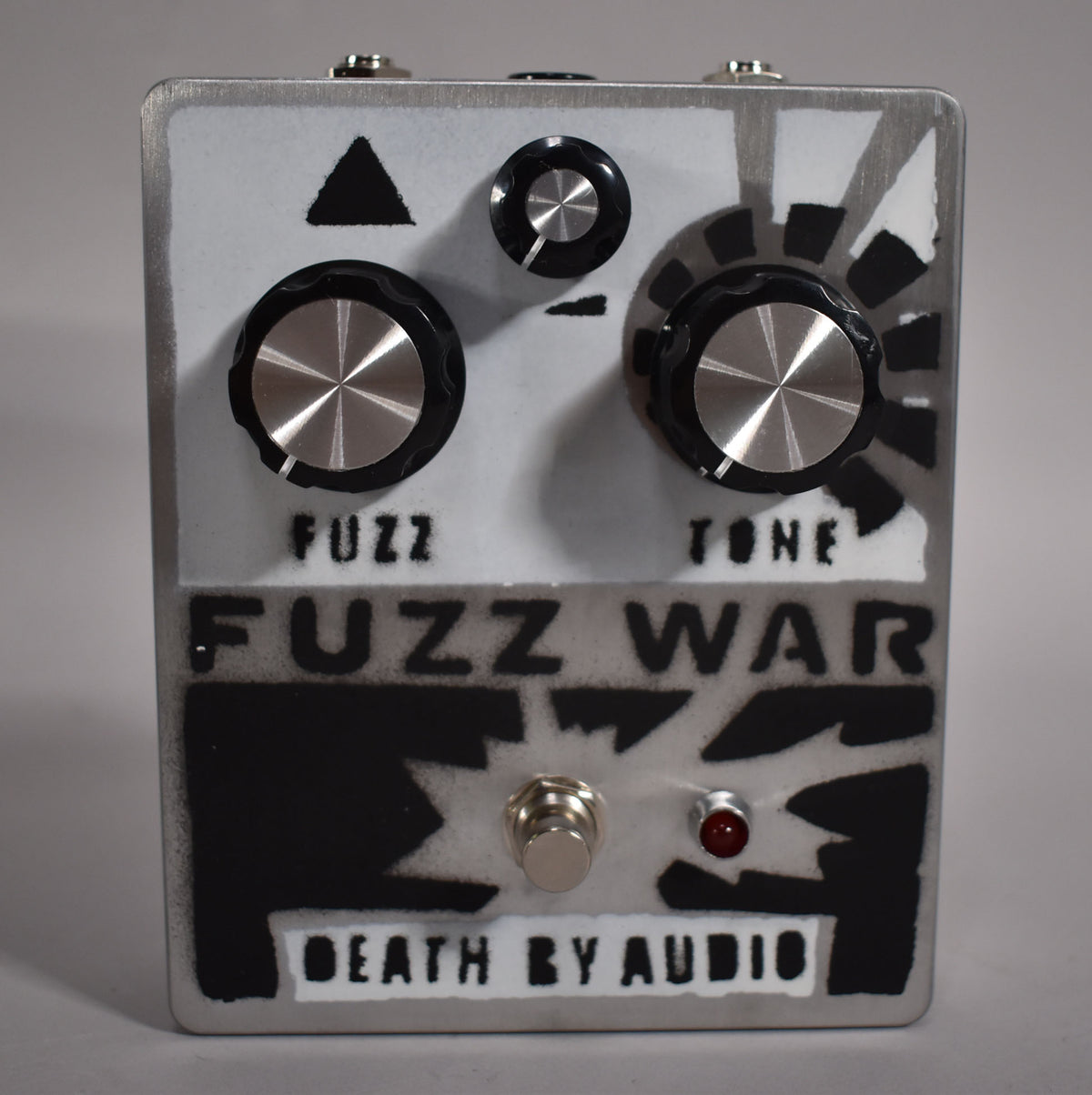 A top-down view of a grey and black fuzz pedal with three black knobs, a footswitch, and a red LED.