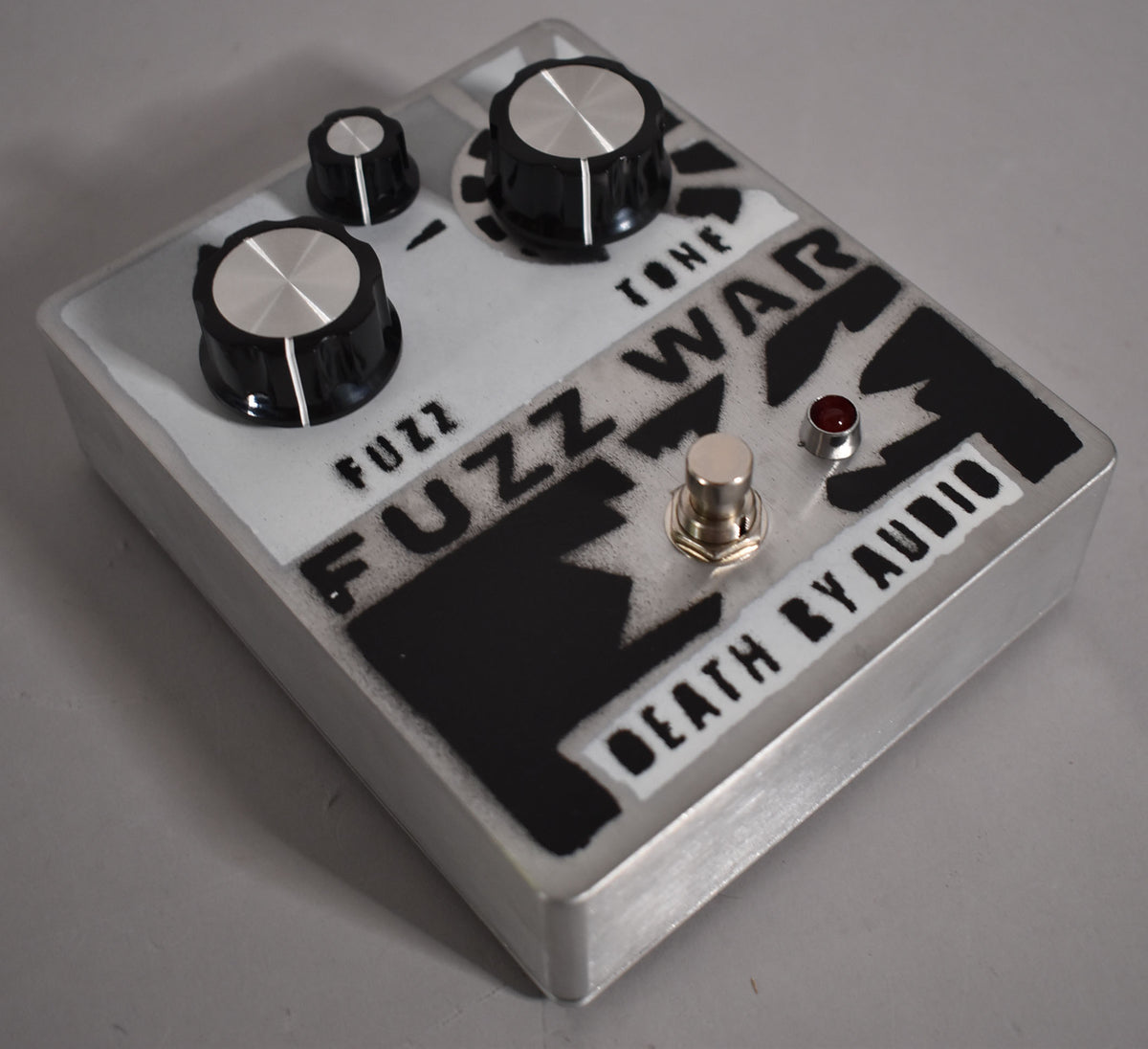 A silver-grey pedal with black stenciled graphics, three black knobs with silver tops, a silver footswitch, and a red LED.