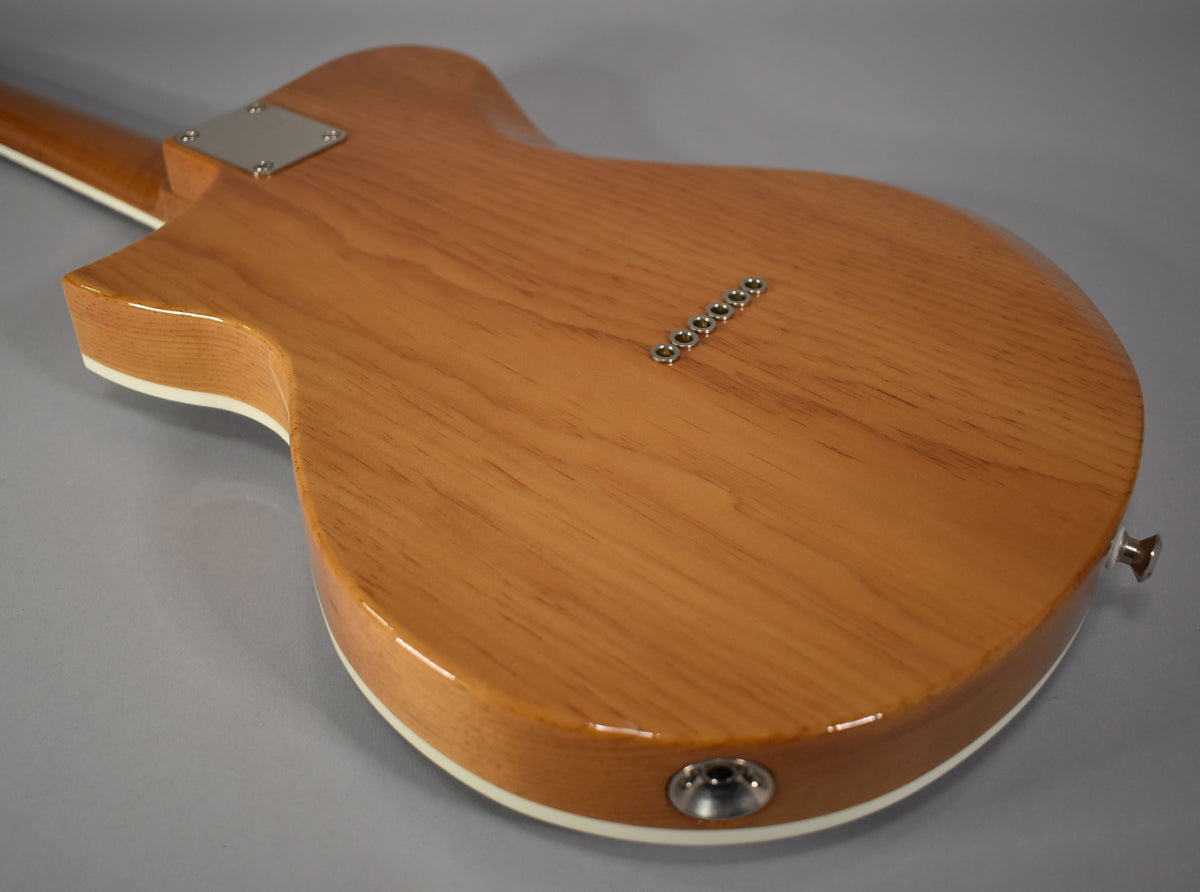 The back of a light brown wooden guitar with a white binding, showing the neck plate, string ferrules, output jack, and strap button.