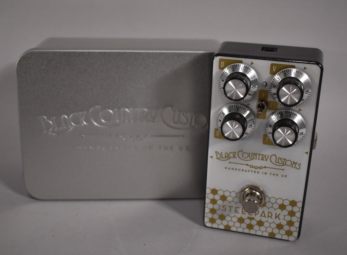 A white pedal with gold honeycomb pattern, four silver-capped black knobs, a toggle switch, and a footswitch, next to a silver tin box.