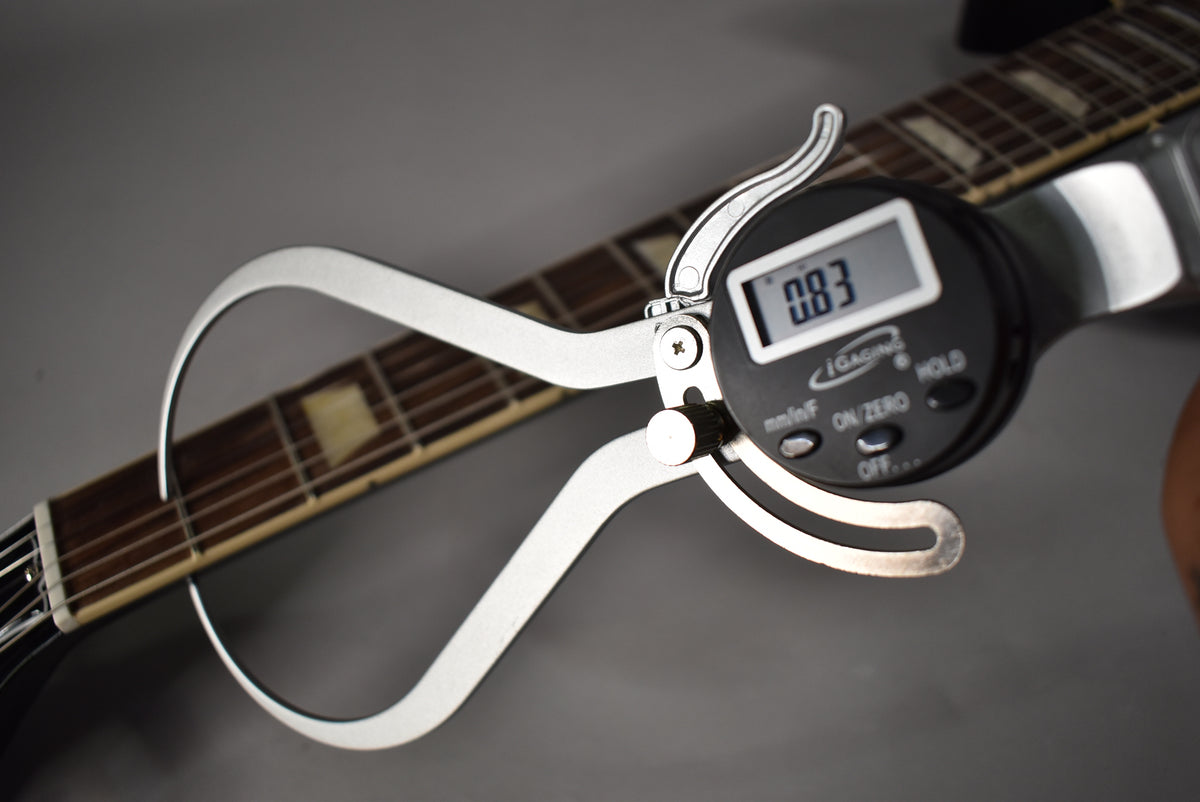A digital caliper measures the string height at the 12th fret of the ebony Les Paul fretboard with trapezoid inlays.