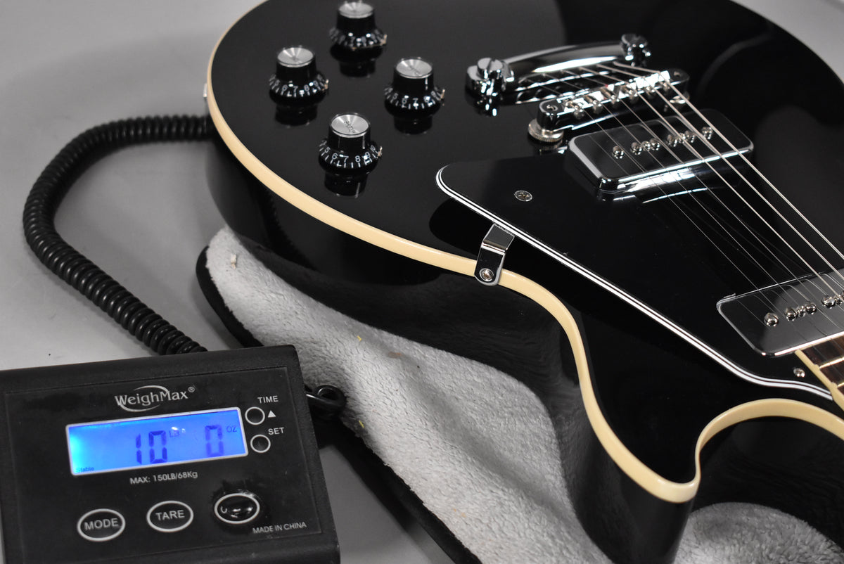 The ebony Les Paul body, with cream binding and chrome hardware, rests in its case next to a scale displaying "10.0" lbs.