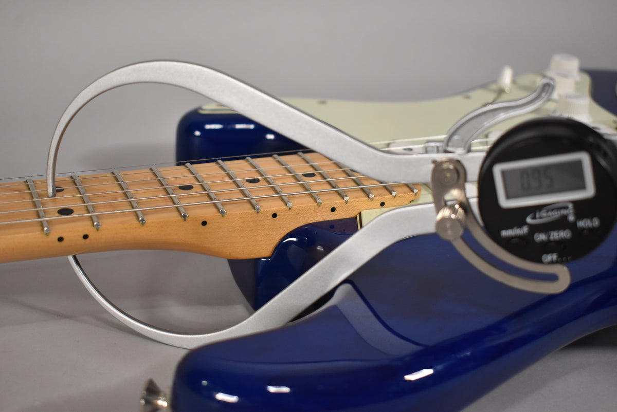 A sapphire blue Stratocaster with a maple fretboard is shown with a digital caliper measuring the neck relief at the 7th fret.