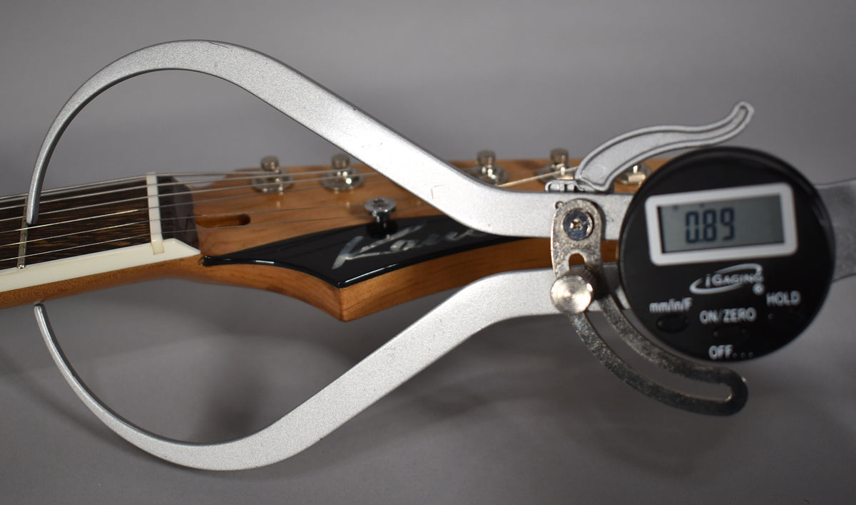 A digital caliper measures the string height at the first fret of the guitar's natural wood neck, showing 0.89mm.