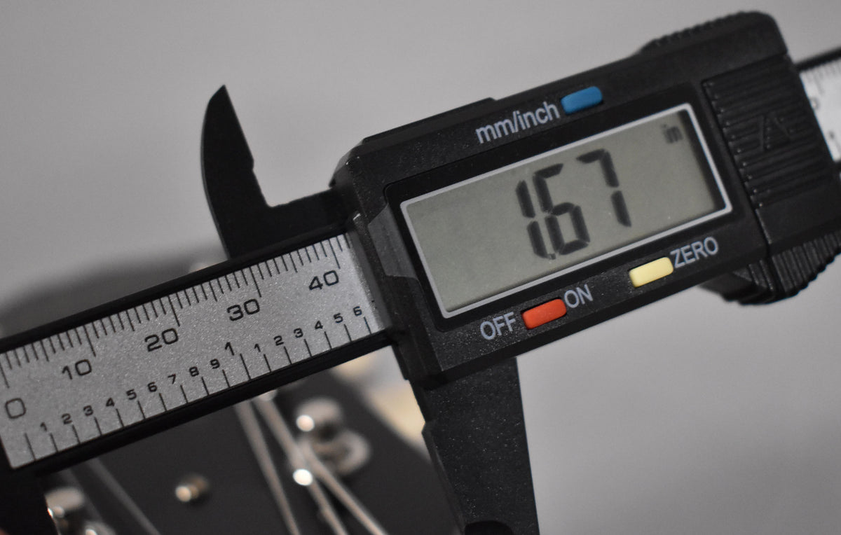 A digital caliper measures the guitar's nut width at 1.67 inches, with tuning pegs and strings visible below.