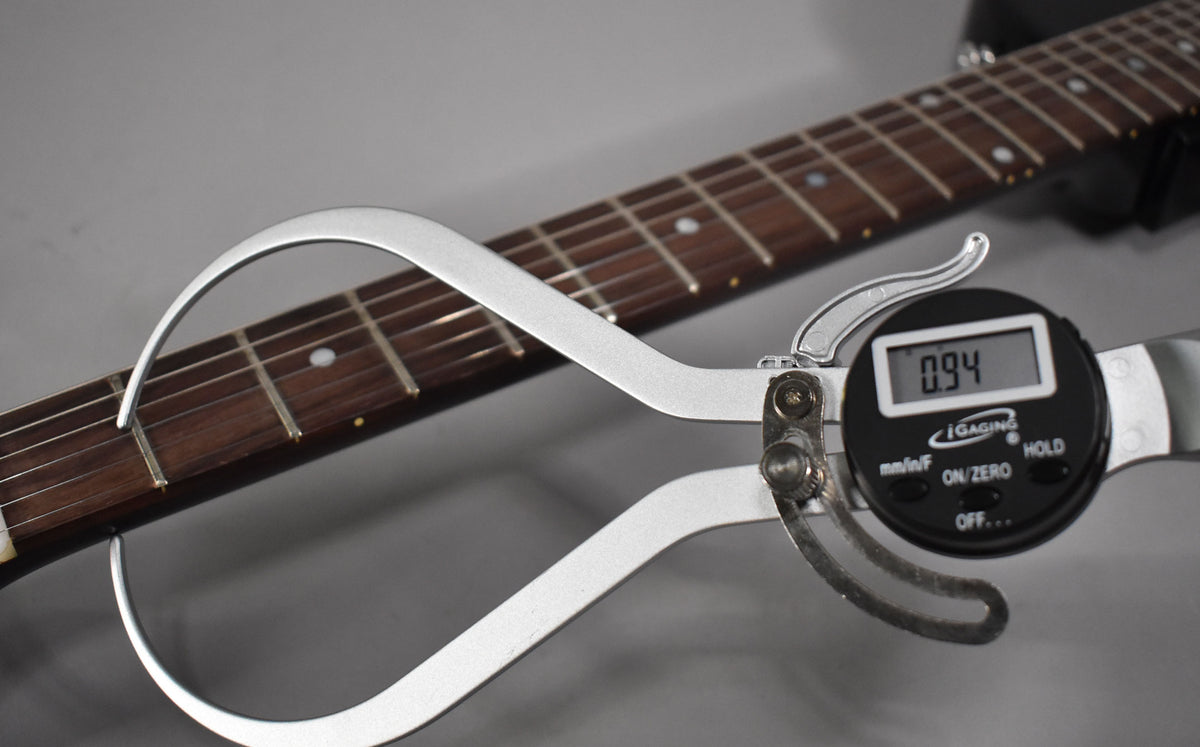 A digital fret caliper measures the guitar's dark rosewood fretboard, showing 0.94 on its display.