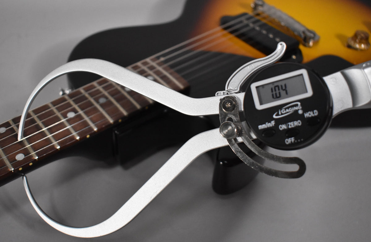 A digital caliper measures the guitar's fretboard radius, showing "1.04" on its display, with the sunburst body blurred behind.