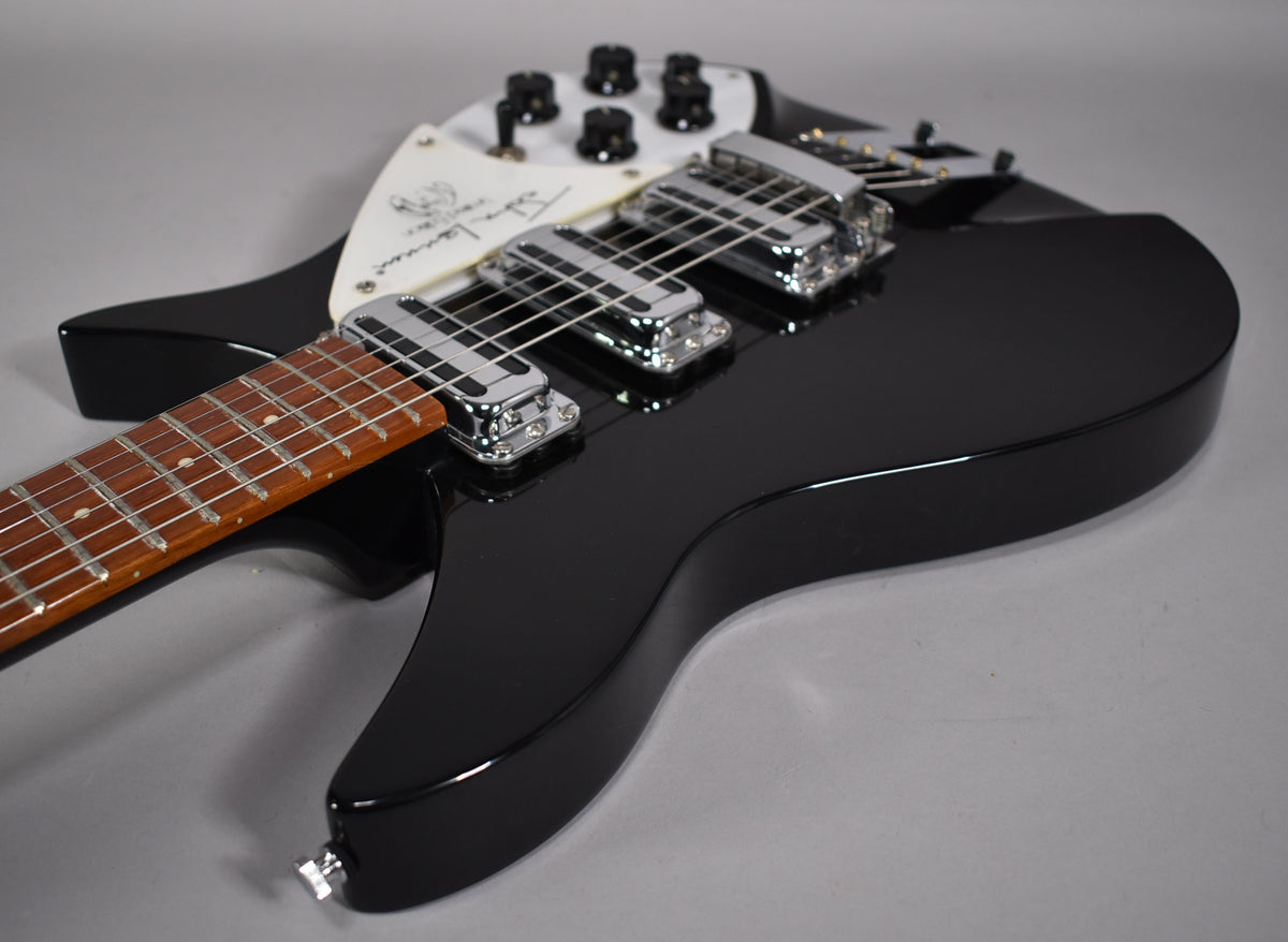A black Rickenbacker guitar with a white pickguard signed "John Lennon" and chrome pickups, angled on a gray surface.