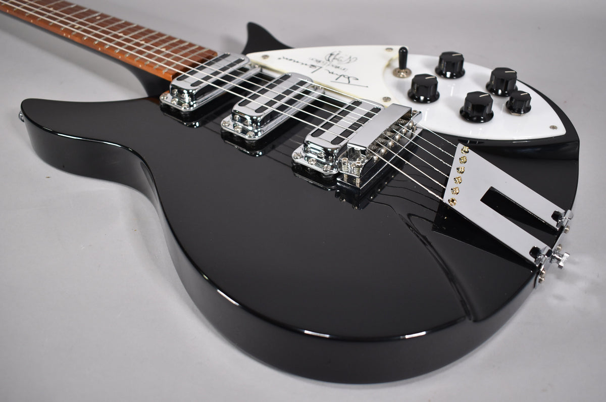 Close-up of a glossy black electric guitar, angled from the lower bout, showing three chrome pickups and a white pickguard with controls.