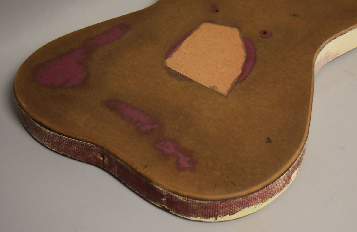 The guitar body's back is angled, showing its brown finish with exposed magenta paint, a routed cavity, and worn red binding.