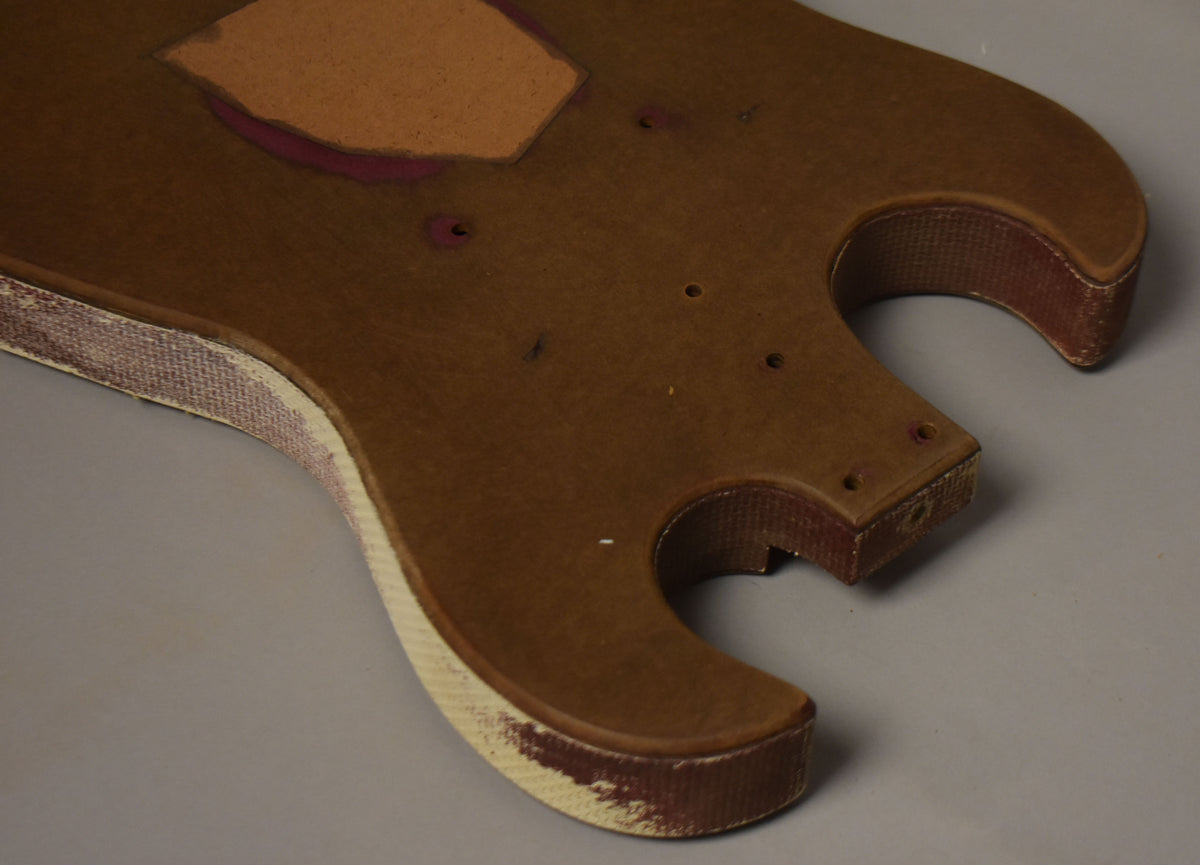 The brown fiberboard guitar body, viewed from above, shows a routed pickup cavity and multiple screw holes, with worn white binding.