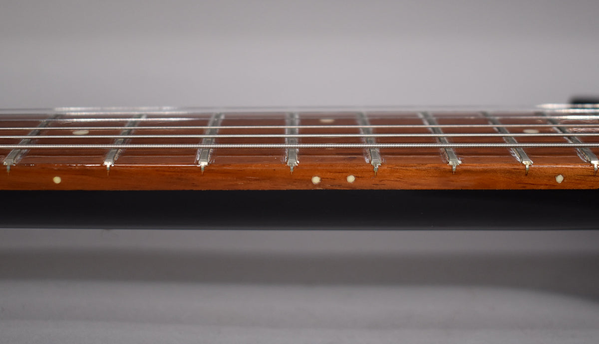 A close-up, side view of a guitar fretboard with strings, frets, and small white dot inlays.