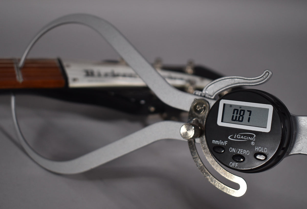 A digital caliper measuring a guitar neck, displaying "0.87" on its screen, with "Rickenbacker" visible on the headstock.