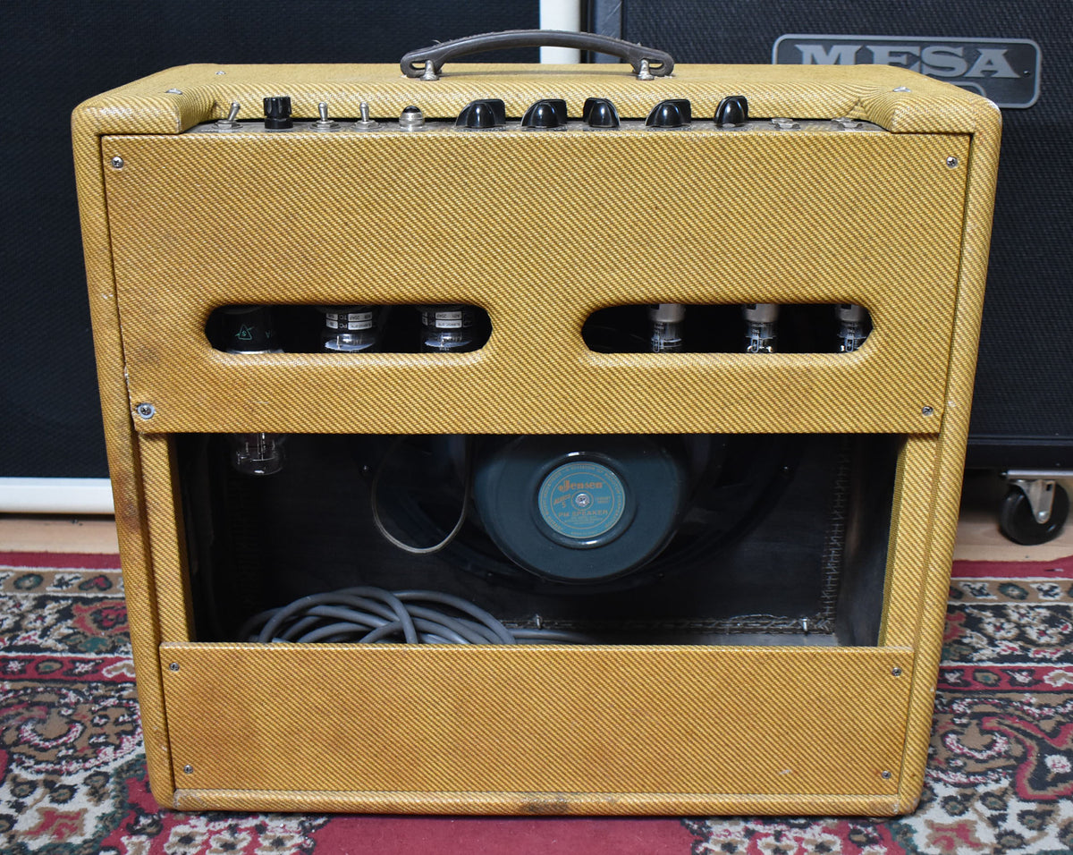 Rear view of a vintage tweed amp, showing the open back with visible tubes, a blue-labeled speaker, and coiled gray cable.
