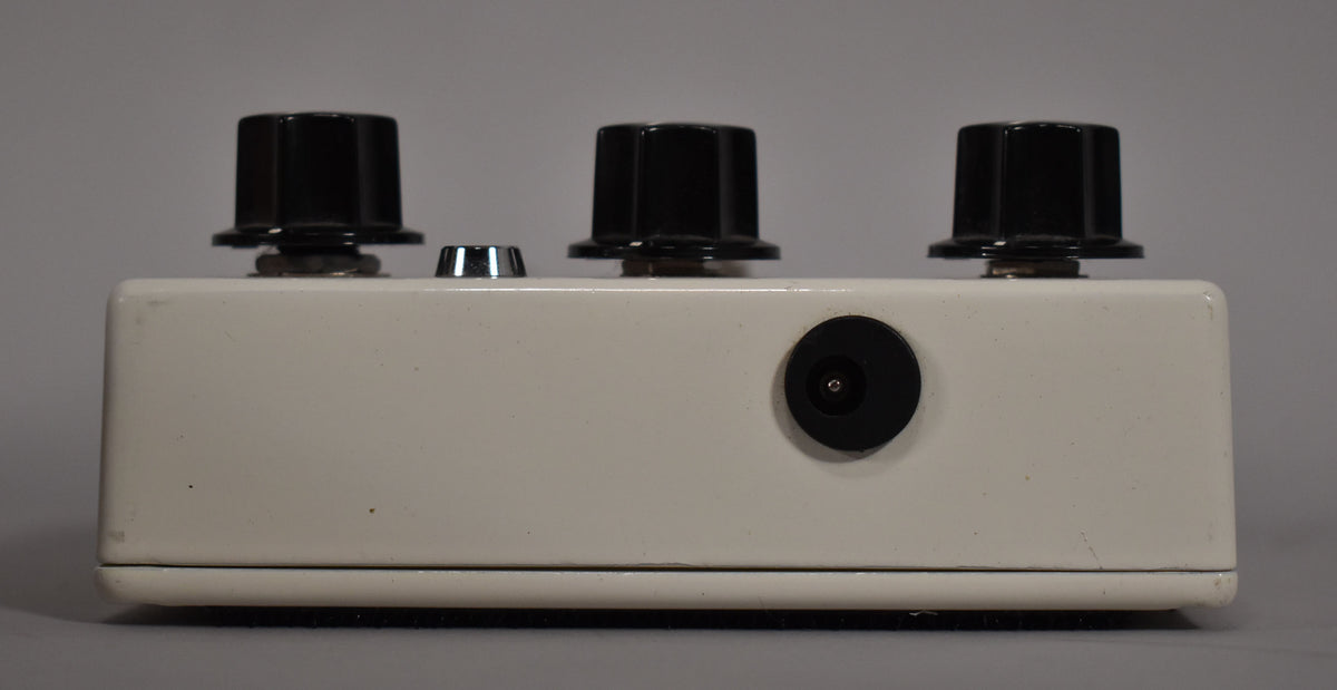 The off-white pedal is viewed from the back, showing a power input and three black control knobs with minor scuffs.