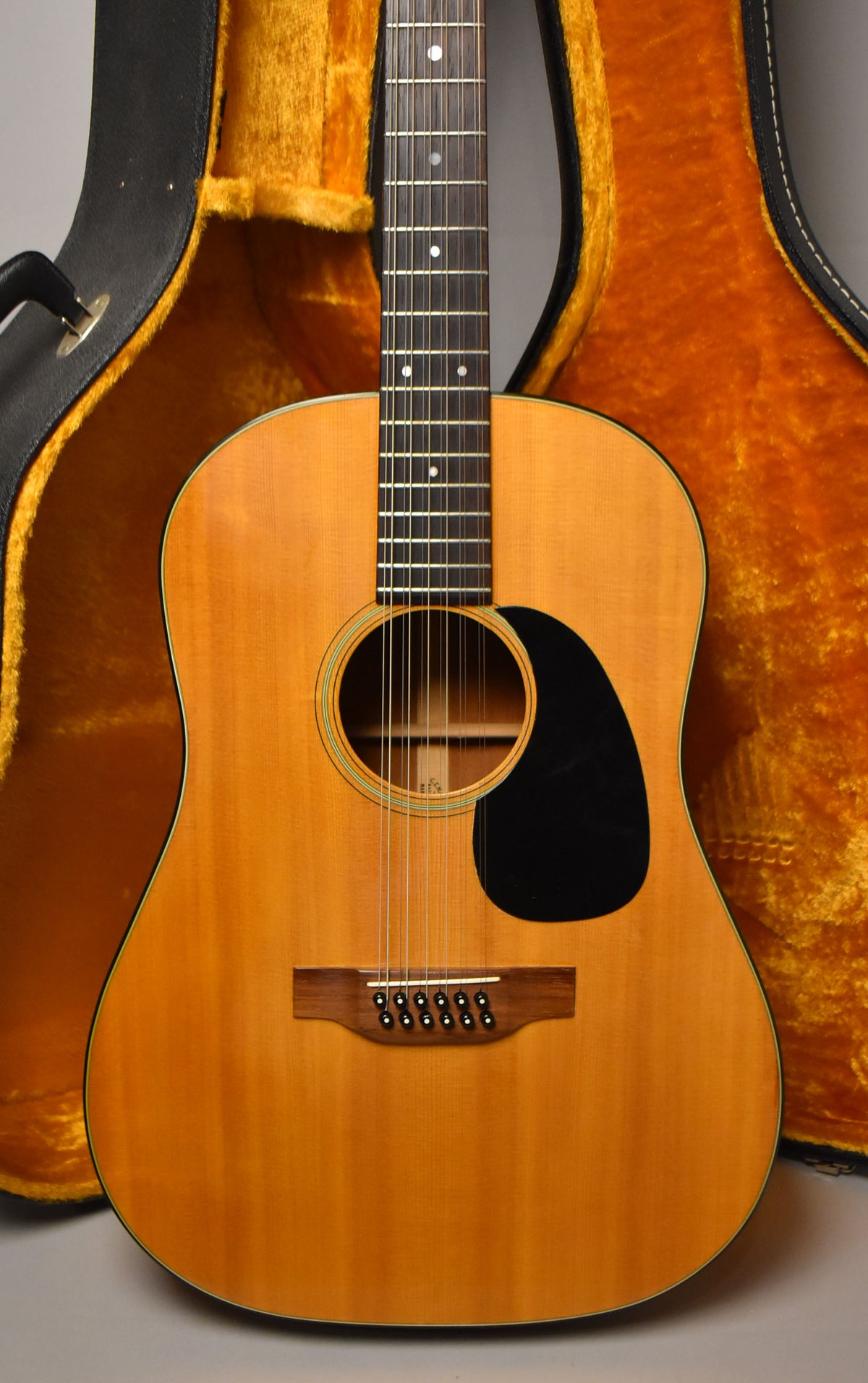 A natural finish 12-string acoustic guitar, with a black pickguard and dark wood bridge, stands in its open, orange-lined hard case.