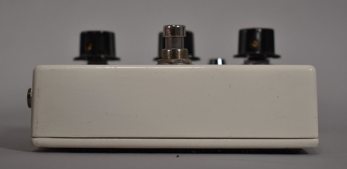 The white pedal is viewed from the side, showing a silver footswitch and two black knobs on top, with a jack on the left.