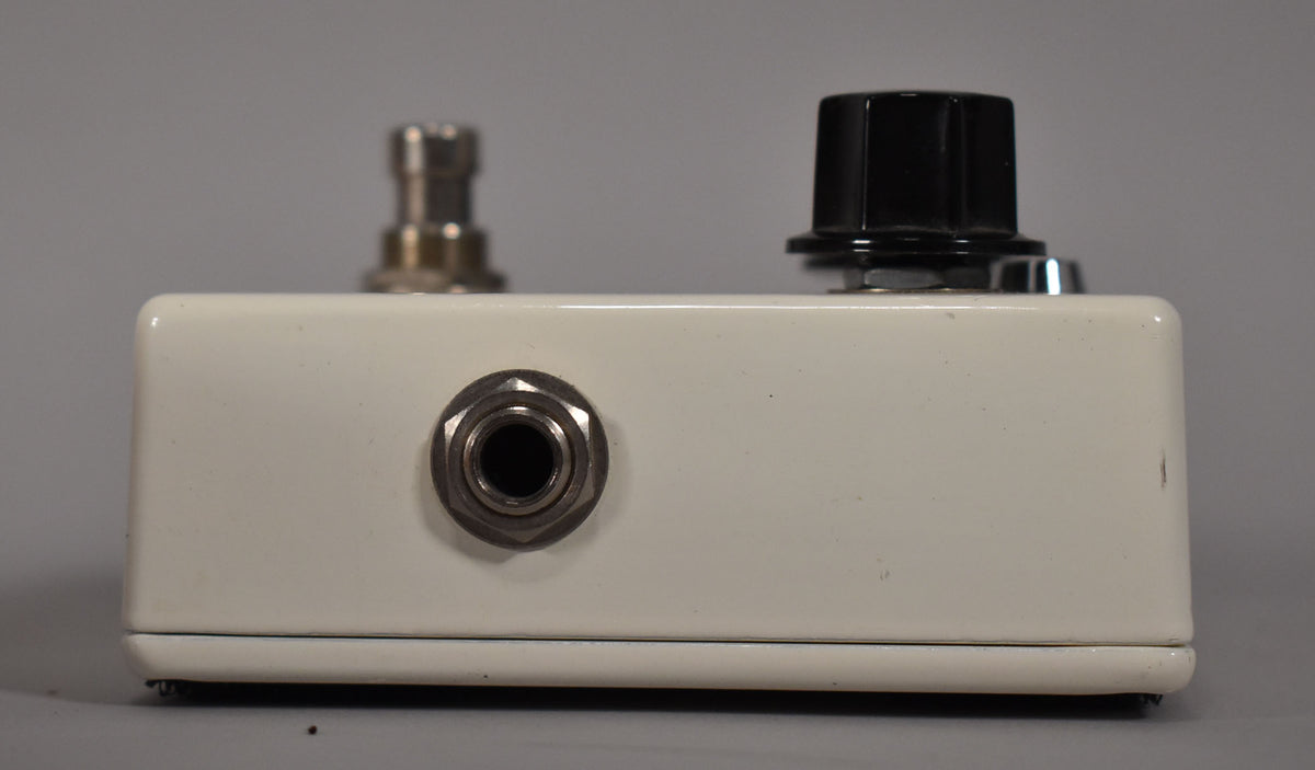 The white pedal is viewed from the side, showing a silver input jack, a black knob, and a silver footswitch.