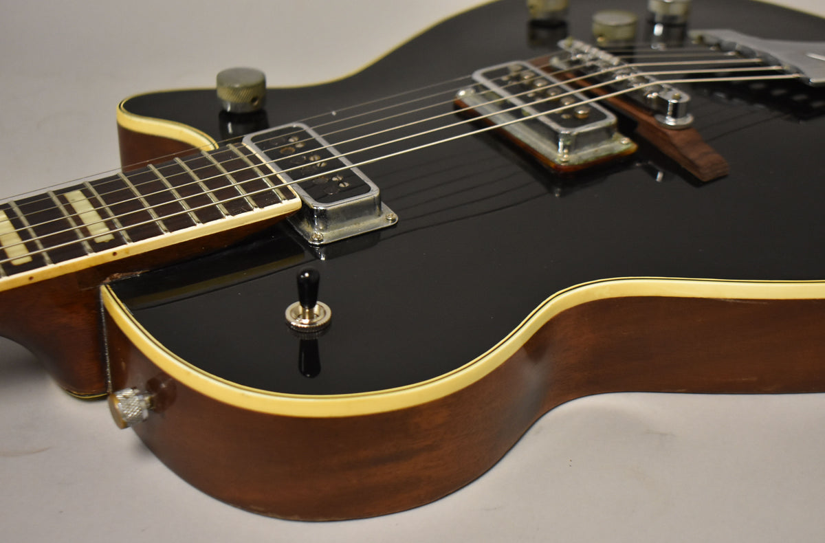 Close-up of a black electric guitar's body and neck, showing two chrome pickups, a black toggle switch, and cream binding.