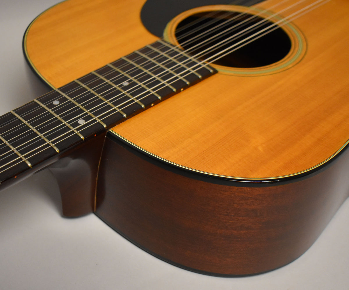 Upper body of a natural finish 12-string acoustic guitar, angled from the neck to the soundhole, showing the dark pickguard and binding.
