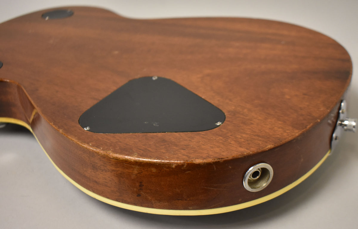 The guitar's dark brown back and side, with a black control plate and chrome output jack, show visible wear and finish checking.