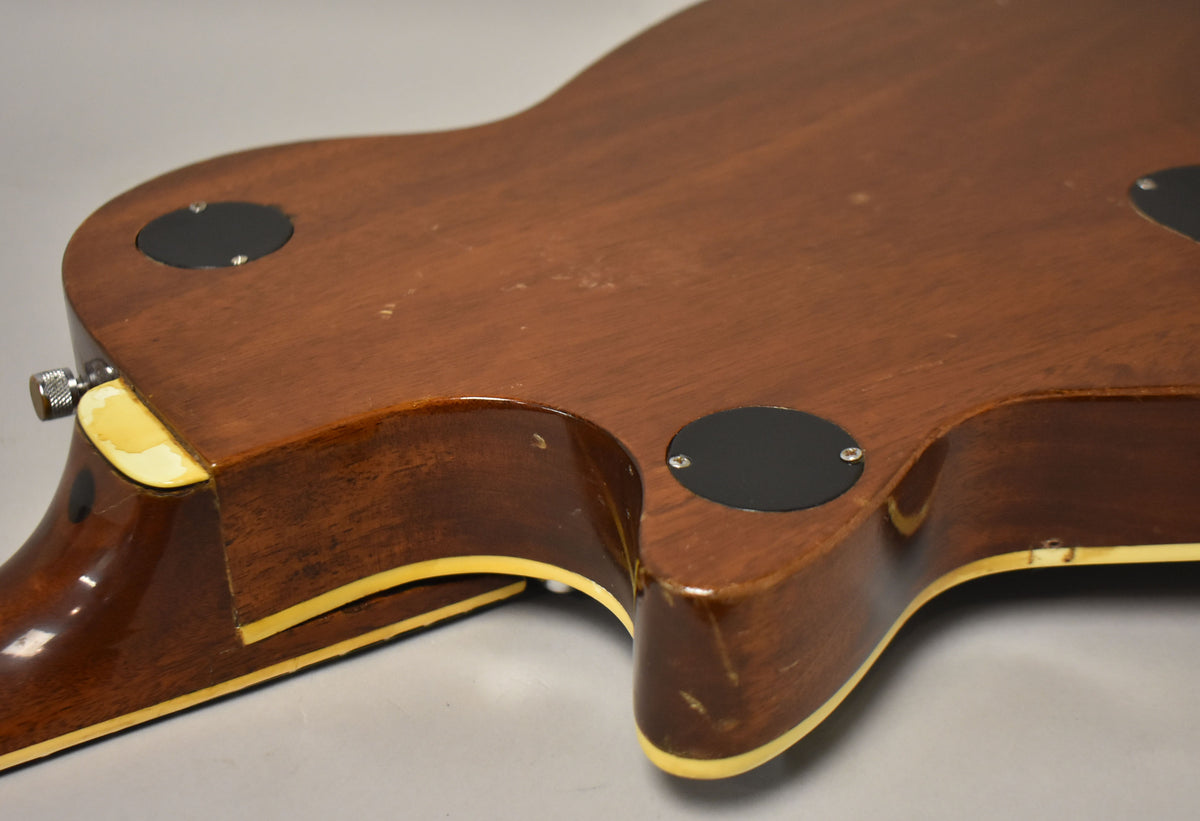 The guitar's dark brown back shows wear, with two black control cavity covers and a chipped cream binding near the neck heel.