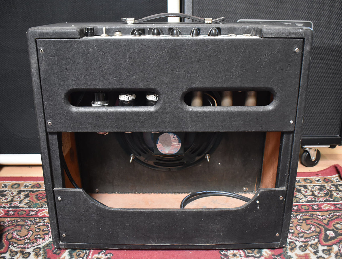 Rear view of a black amp with a large speaker visible through the open back, showing tubes and controls on top.