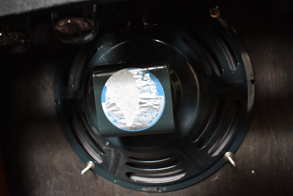 Close-up of the amp's dark speaker, showing its center magnet with a partially peeled silver and blue sticker, and two vacuum tubes.