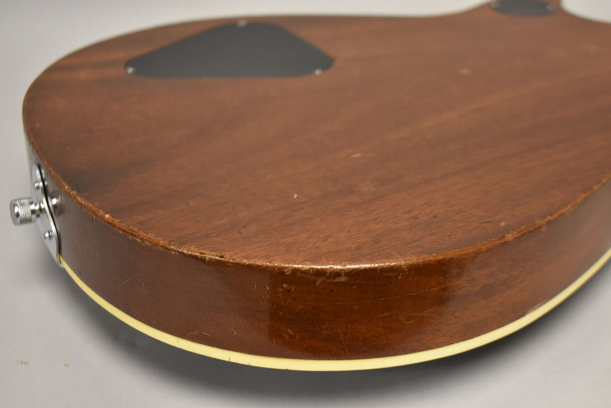 The guitar's dark brown back and side are visible, with a chrome output jack and binding, plus wear and checking along the edges.