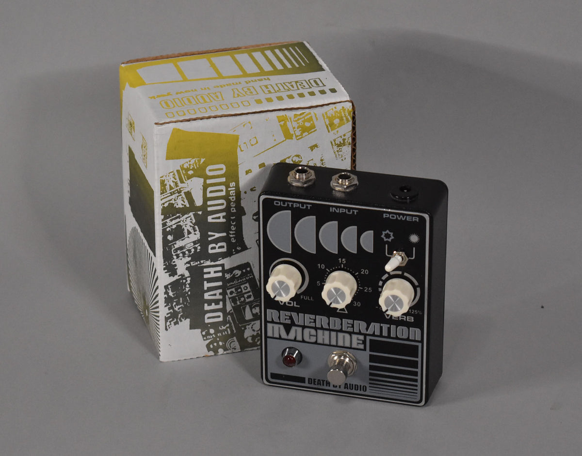 Black pedal with white knobs and graphics, viewed from the front-right, next to its partially visible cardboard box.