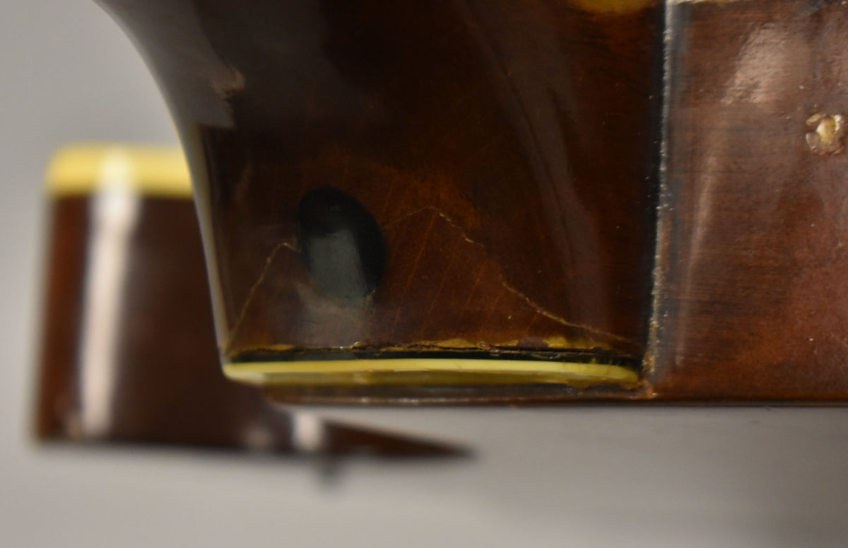 Close-up of the guitar's dark brown neck heel, showing finish checking, a black strap button, and cream binding with a thin black line.