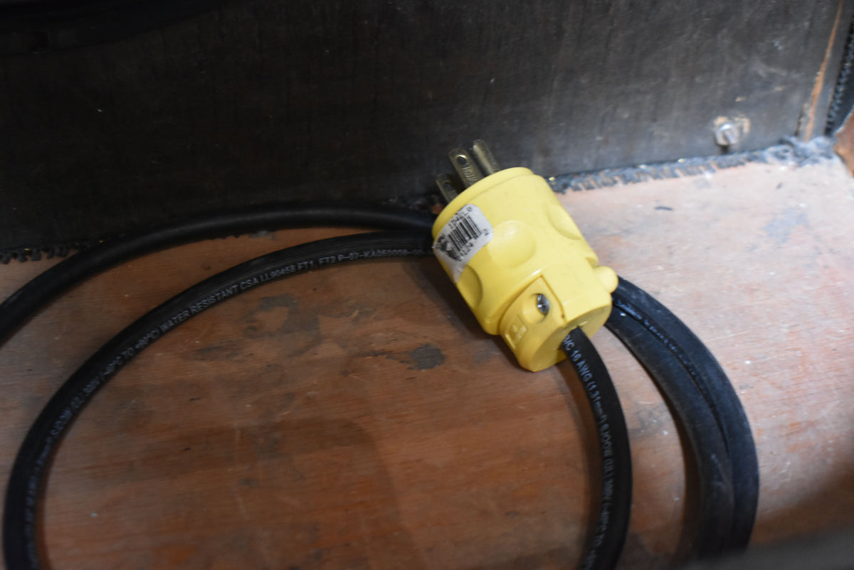 A yellow three-prong power plug with a black cord coiled on a wooden surface, with a dark amp cabinet visible in the background.