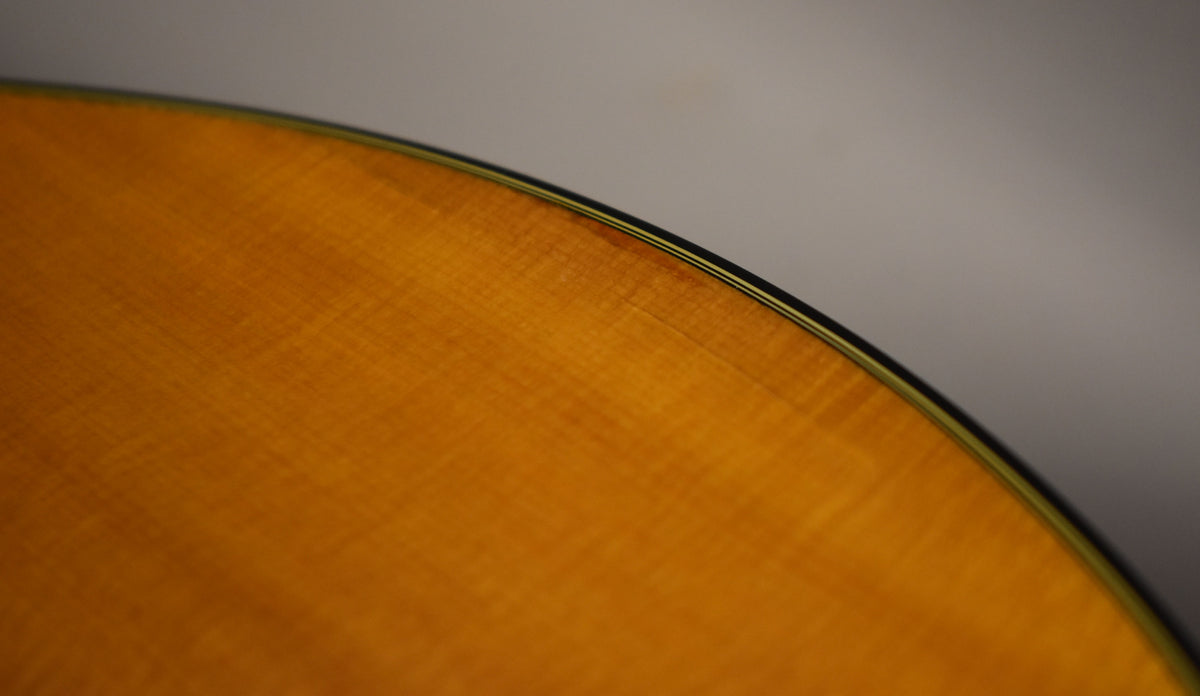 Close-up of the guitar's natural finish top, showing fine grain, light checking, and a dark binding with a thin green stripe.