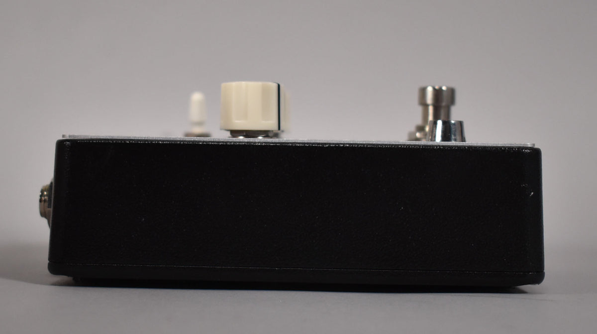 Side view of a black pedal with a silver top plate, showing a cream knob, a small white switch, and a silver footswitch.