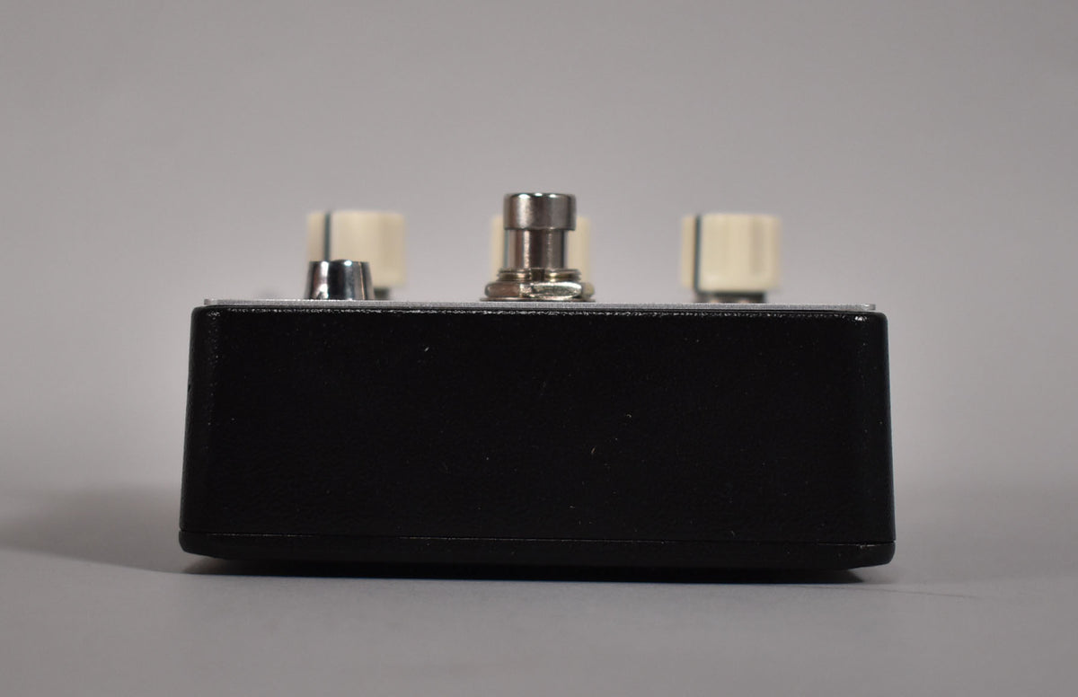 The black pedal is viewed from the side, showing a silver footswitch and two cream control knobs on top.