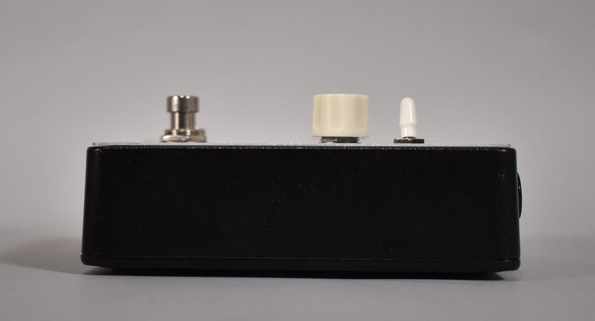 Side view of the black textured pedal with a silver top, showing a metal footswitch, a cream knob, and a white toggle switch.