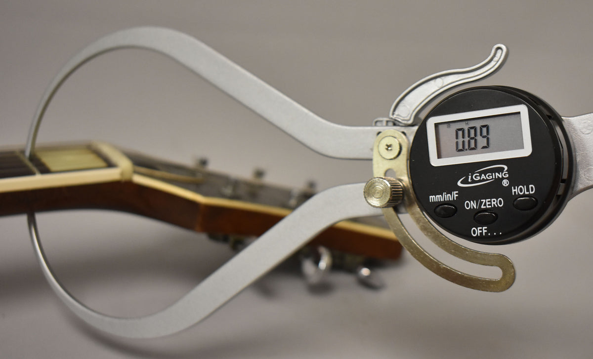 A digital caliper measures the guitar's nut, showing "0.89" on its display, with the headstock blurred in the background.