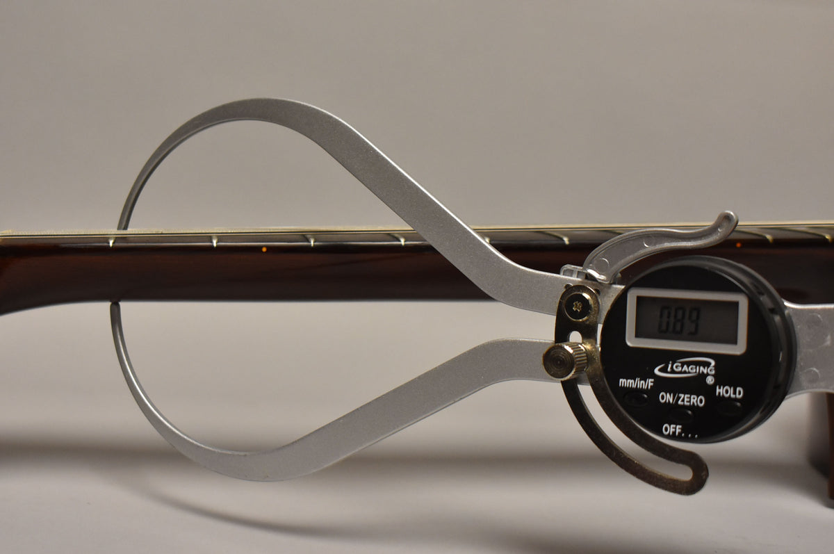 A digital caliper measures the guitar's dark brown neck and fretboard, showing a reading of 0.089.