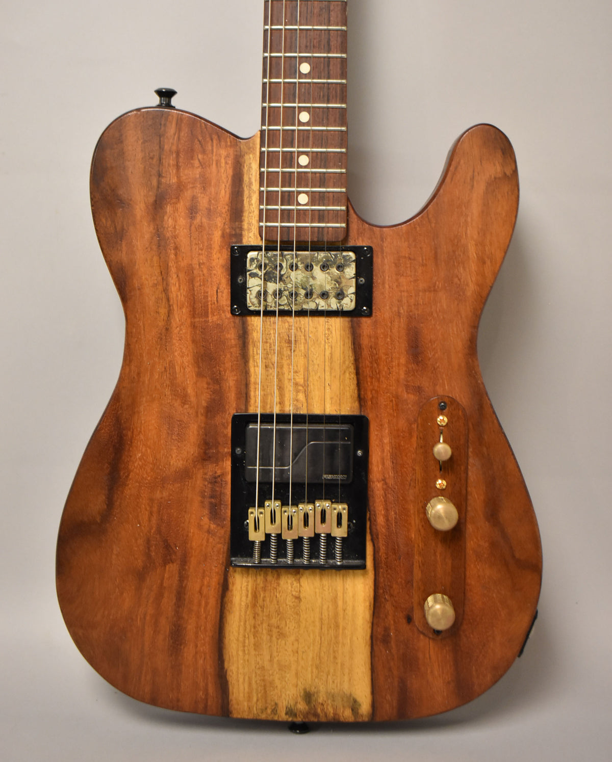 Close-up of a natural finish T-style guitar, showing the body's two-tone wood grain, two humbuckers, and brass control knobs.