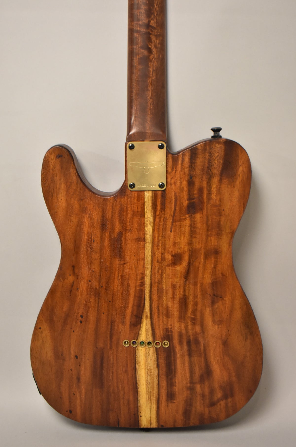The back of a T-style guitar, showing a natural dark wood body with a lighter wood stripe, brass neck plate, and six brass string ferrules.