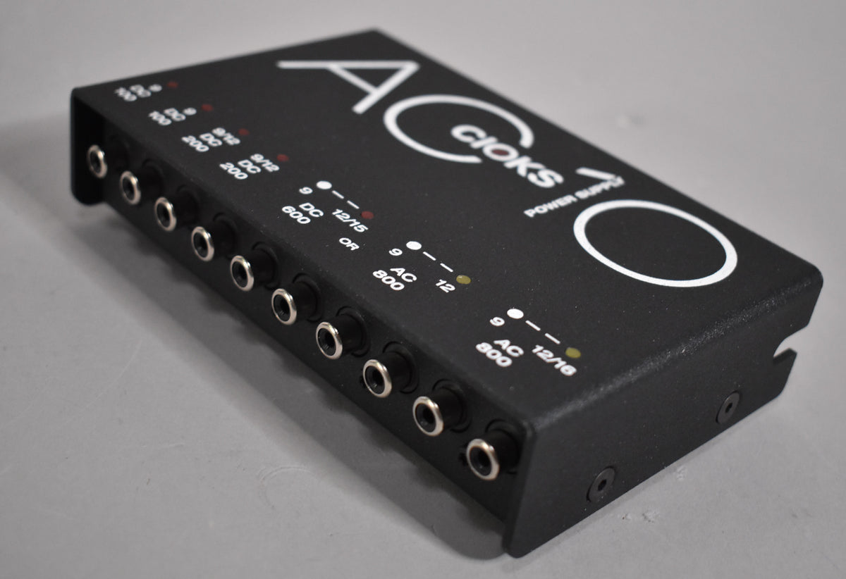 A black textured power supply with 10 silver-ringed output jacks, viewed from the front-left, showing white text and red/yellow indicator lights.