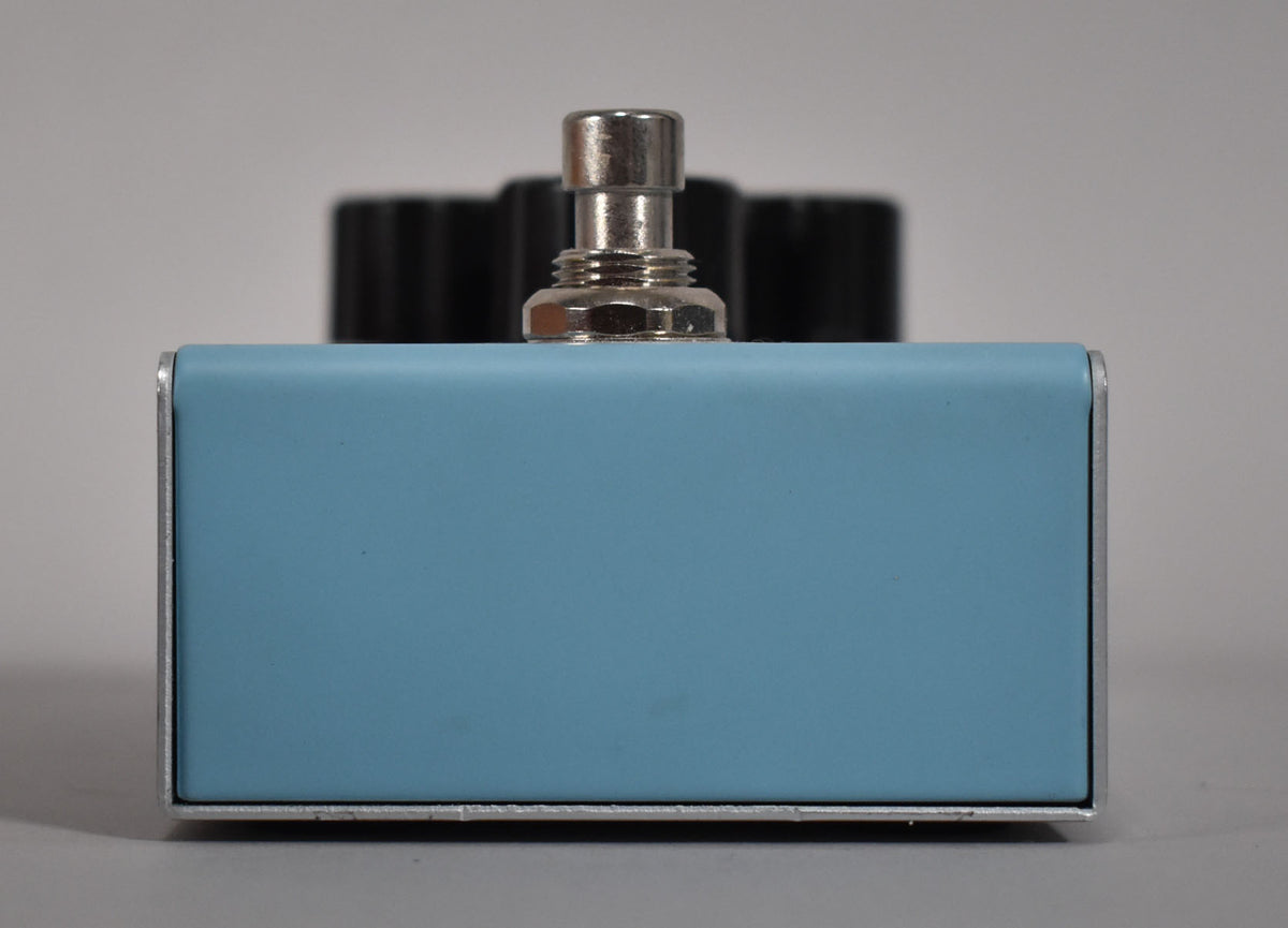 The light blue pedal is viewed from the front, showing a chrome footswitch and three black control knobs blurred in the background.