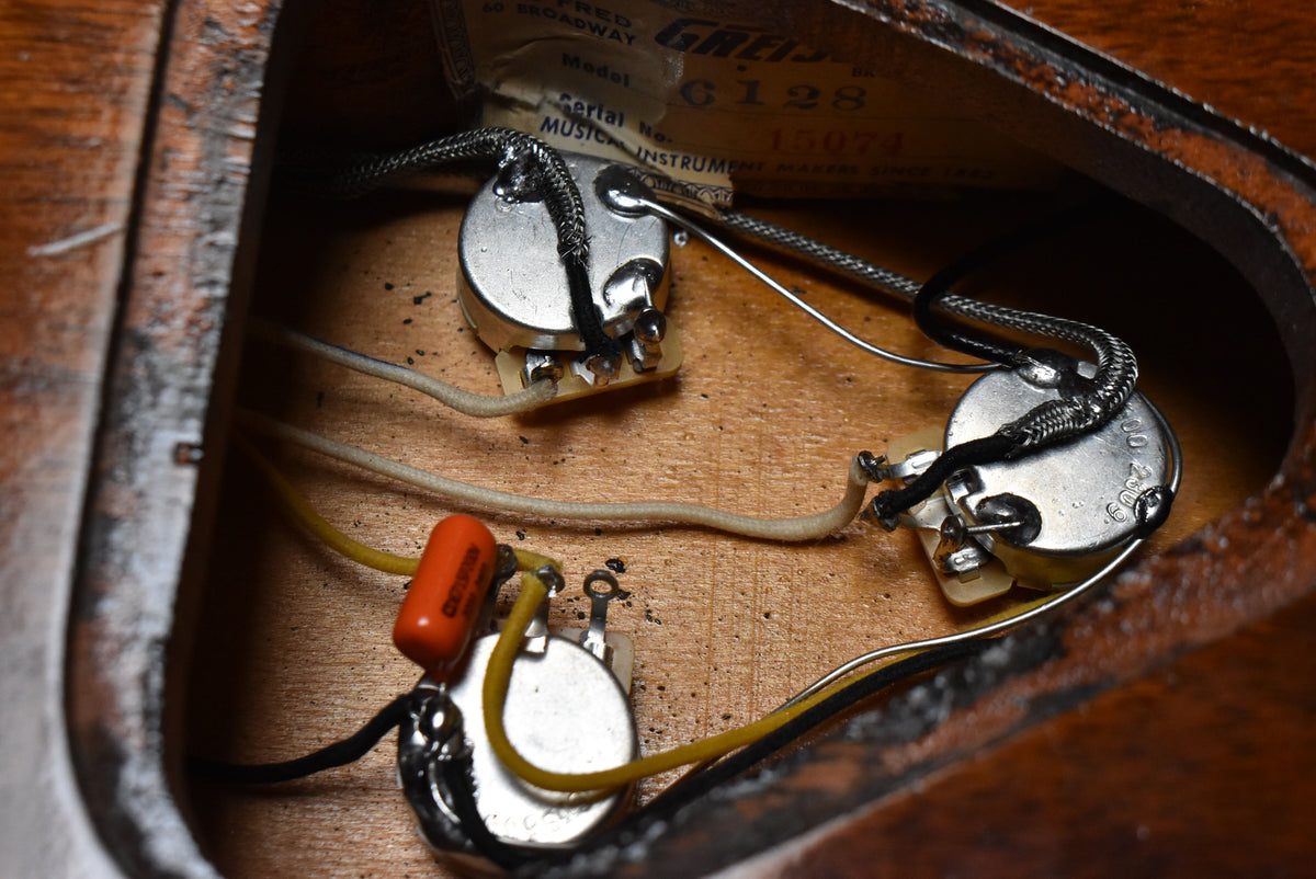 Close-up of the guitar's internal wiring, showing three silver potentiometers, an orange capacitor, and various wires on a wooden surface.