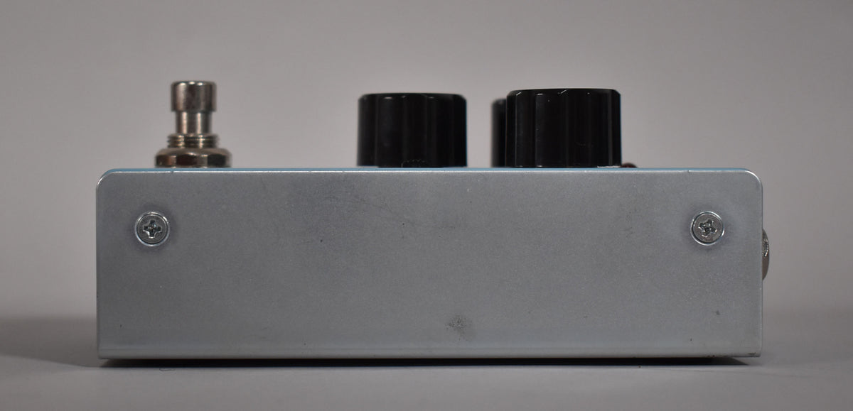 The reverb pedal is viewed from the side, showing its light blue top, silver-grey metal casing, and three black control knobs.