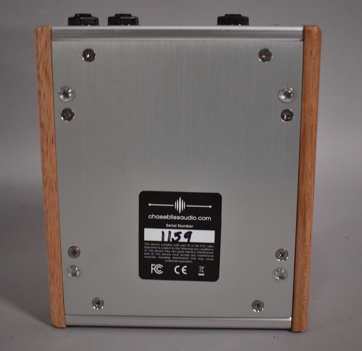 The pedal's back panel is brushed silver metal with light wood sides, showing a black serial number sticker and eight visible screws.