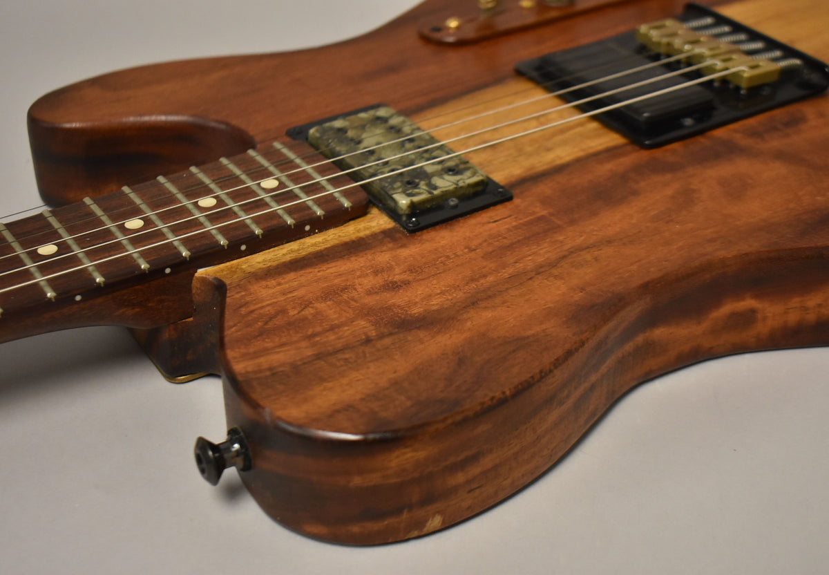Close-up of a natural finish guitar body and neck, showing two pickups, strings, frets, and an output jack.