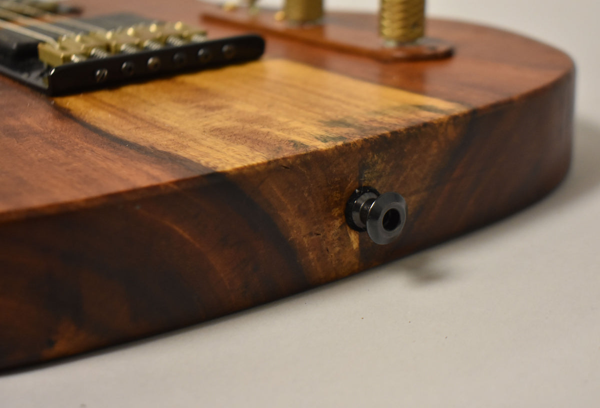 Close-up of the guitar's lower bout, showing the natural wood grain, dark output jack, and part of the black bridge with brass saddles.