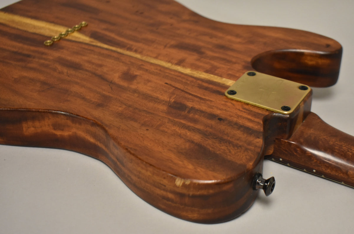 The back of a natural finish T-style guitar, showing a brass neck plate, string ferrules, and a chip on the lower bout.